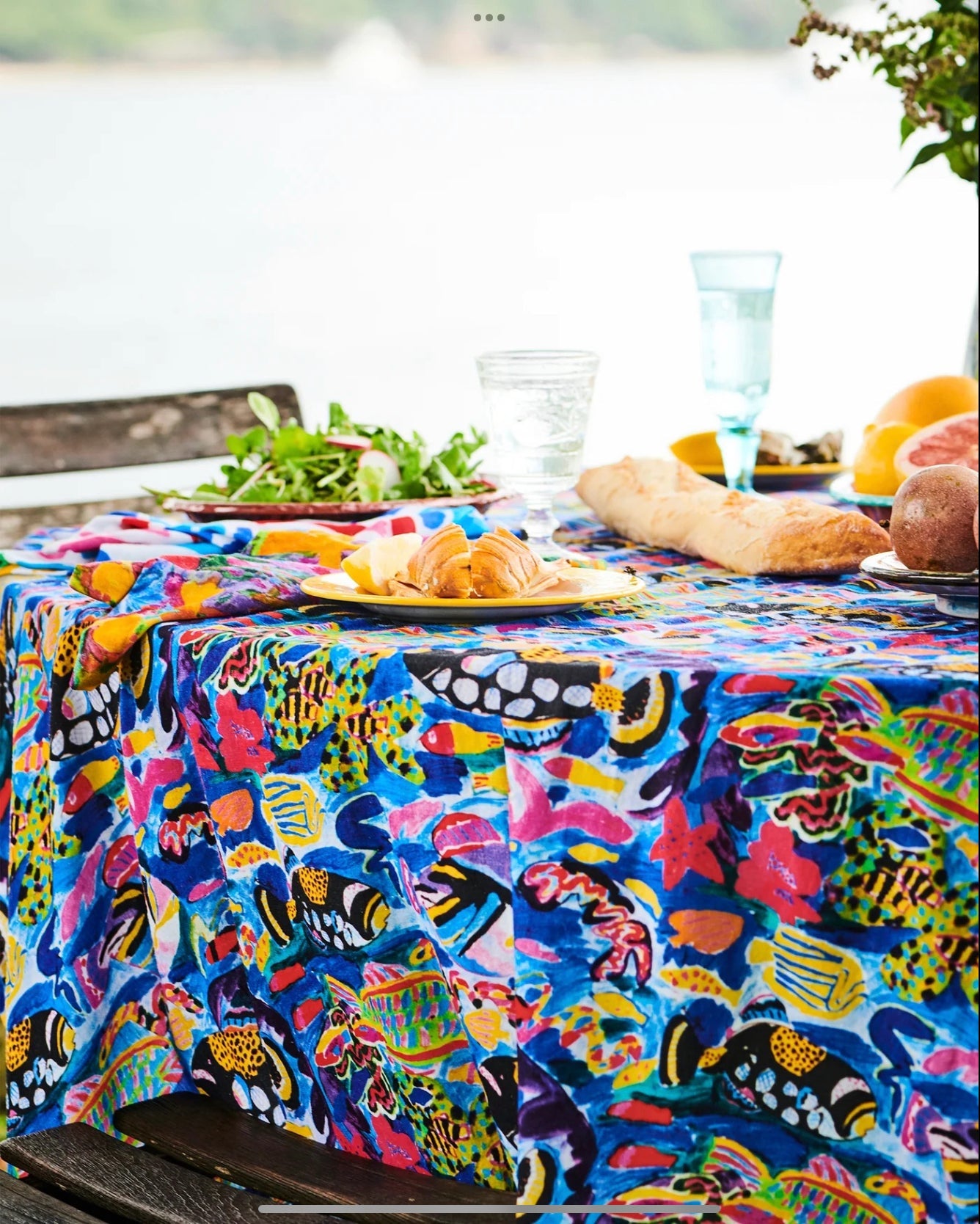A round French flax linen tablecloth by Kip and co featuring ken done artwork of the Great Barrier Reef. Table is set for a casual lunch by the water. 